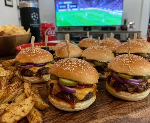Mini hamburguesas BBQ con queso cheddar gratinado, pepinillos y cebolla morada atravesadas con palillo, servidas con papas en gajos para ver fútbol