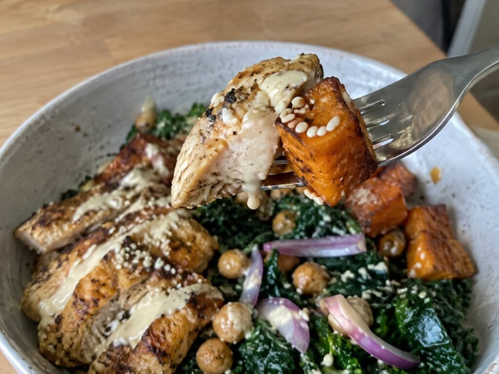Tenedor con un bocado de pollo jugoso y camote asado bañado en aderezo de tahini y limón, bowl saludable alto en proteína