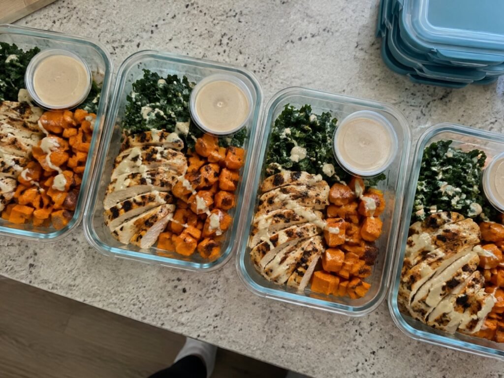 Cuatro tuppers de vidrio con meal prep de pollo, camote asado y kale con aderezo de tahini aparte, comidas saludables para la semana