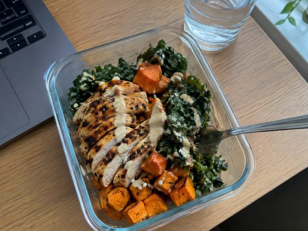 Tupper con bowl de pollo, camote asado y kale con aderezo de tahini en un escritorio junto a la laptop, comida saludable para la oficina