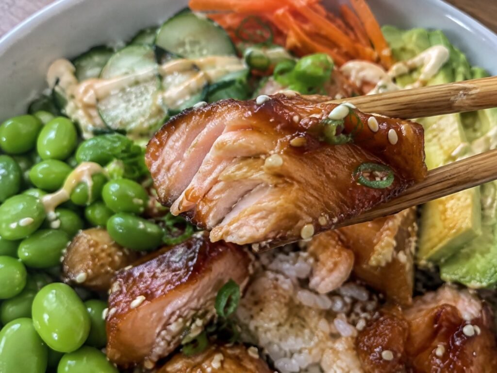 Palillos levantando un trozo de salmón teriyaki jugoso y glaseado sobre bowl de arroz con edamame, receta saludable alta en proteína