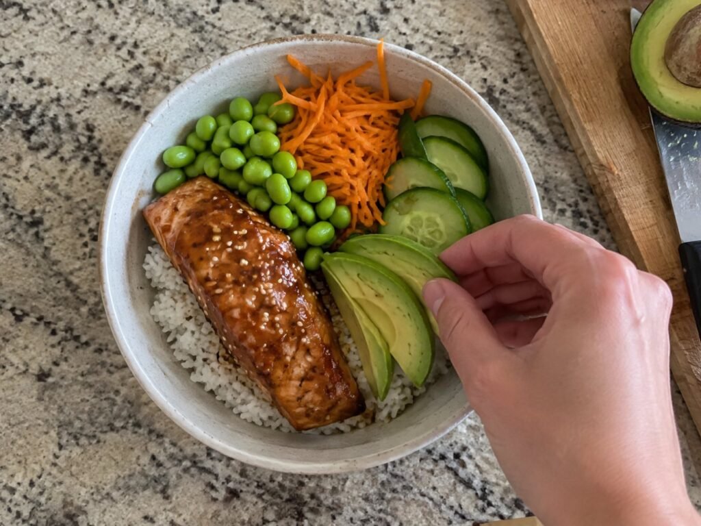 Armando un bowl saludable de arroz con edamame, pepino y zanahoria mientras una mano agrega rebanadas de aguacate en la barra de cocina