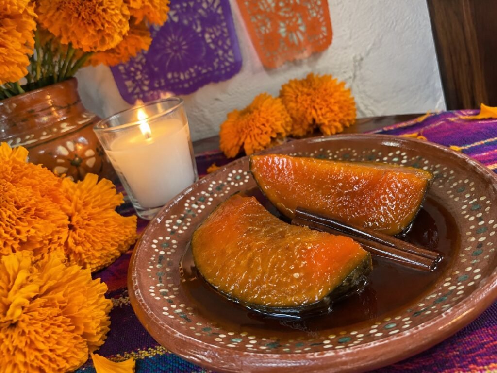 Plato de barro con gajos de calabaza en tacha en almíbar sobre una ofrenda casera de Día de Muertos con flores de cempasúchil y vela