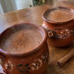 Dos jarritos de barro con champurrado ligero de avena y cacao humeante, espeso y con canela, sobre una mesa de madera en casa