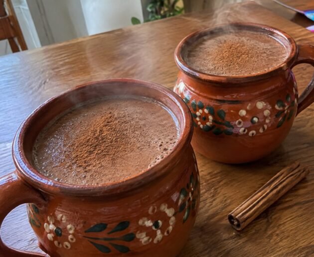 Dos jarritos de barro con champurrado ligero de avena y cacao humeante, espeso y con canela, sobre una mesa de madera en casa