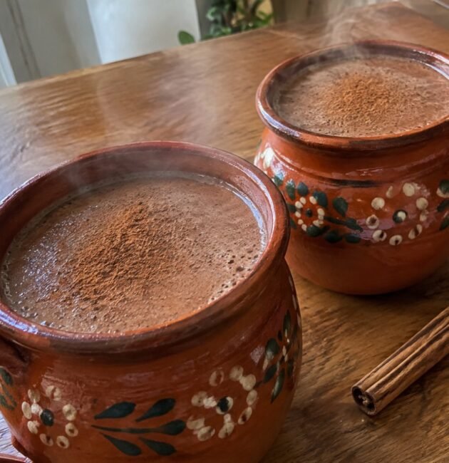 Dos jarritos de barro con champurrado ligero de avena y cacao humeante, espeso y con canela, sobre una mesa de madera en casa