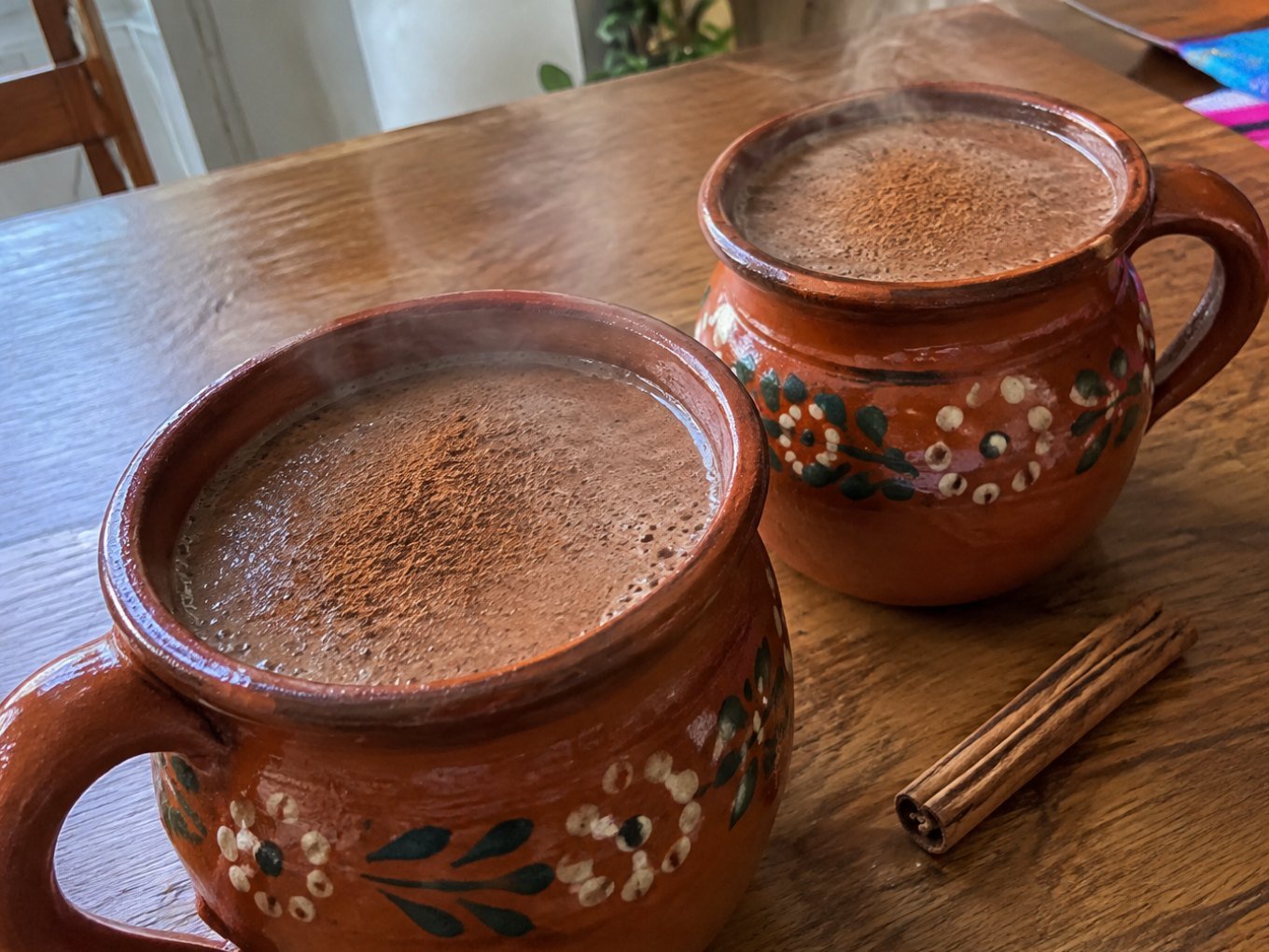 Dos jarritos de barro con champurrado ligero de avena y cacao humeante, espeso y con canela, sobre una mesa de madera en casa