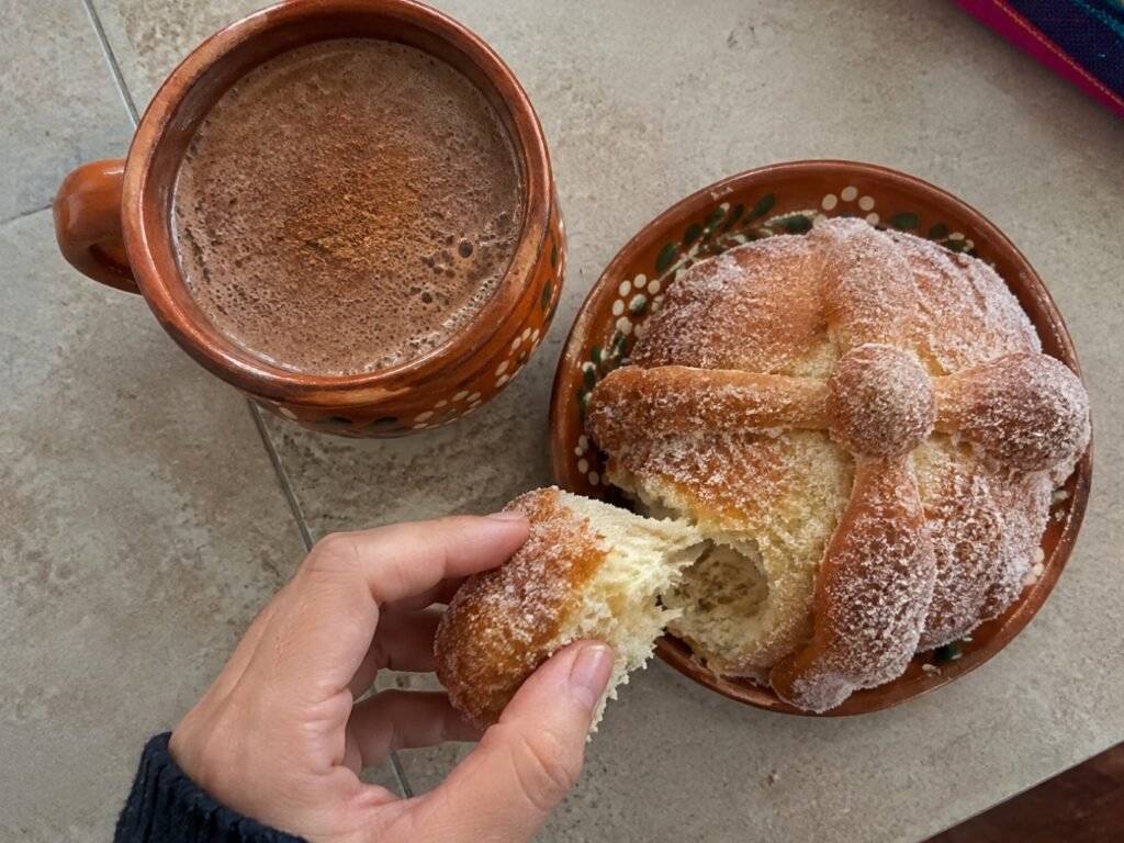 Taza de champurrado de avena y cacao junto a un pan de muerto con azúcar, mano partiendo un pedazo de pan sobre la barra de cocina