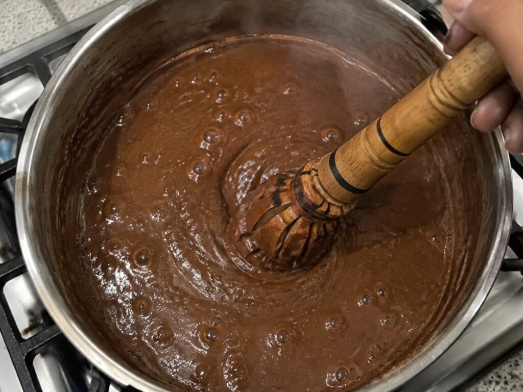 Mano batiendo champurrado espeso de avena y chocolate con molinillo de madera en una olla sobre la estufa de casa, con burbujas