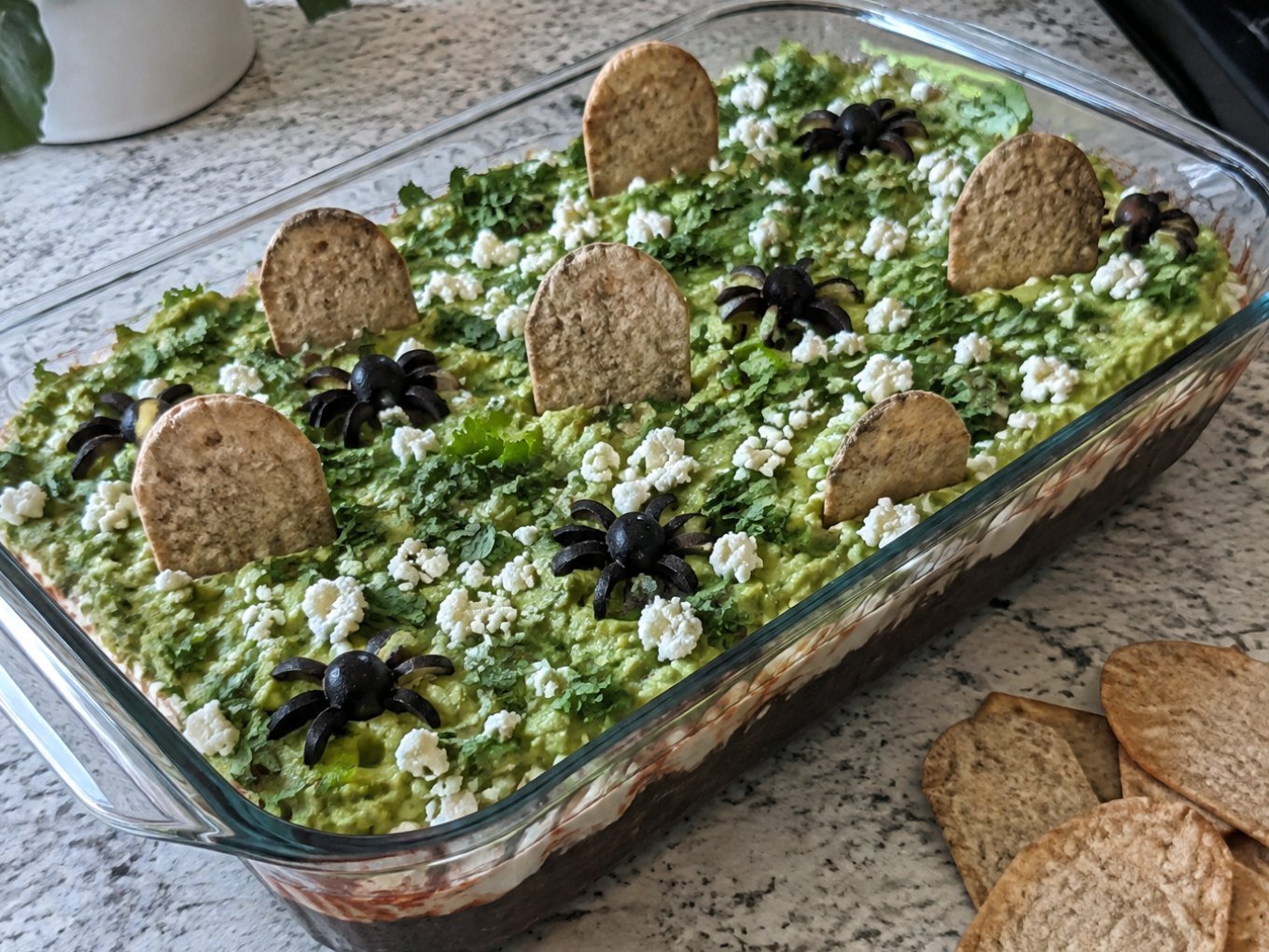 Dip de frijol negro en capas con guacamole, yogur al chipotle y totopos horneados en forma de lápida, botana de Halloween saludable