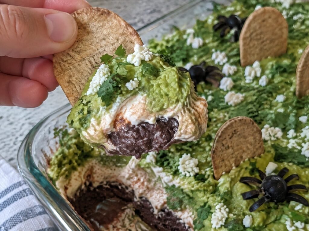 Mano levantando un totopo horneado cargado de dip de frijol negro, yogur griego al chipotle y guacamole, botana mexicana de Halloween