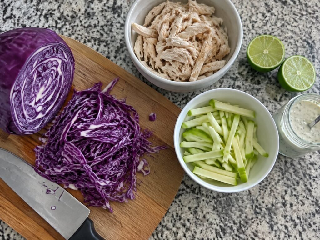 Col morada fileteada en tabla de madera, manzana verde en tiras, pollo deshebrado, limón y aderezo de yogur sobre la barra de la cocina