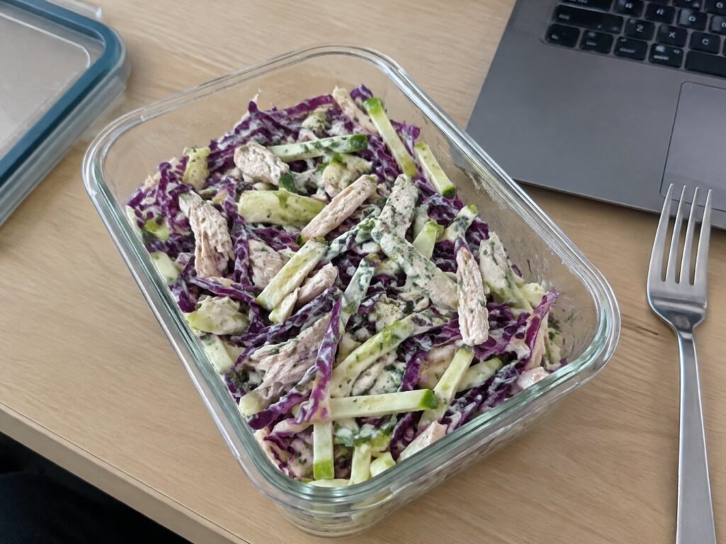 Ensalada de col morada, manzana y pollo con aderezo de yogur en recipiente de vidrio para meal prep sobre escritorio de oficina