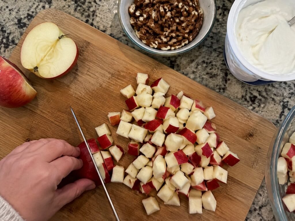 Manos picando manzana roja en cubos sobre tabla de madera con nuez y yogur griego para la ensalada de manzana navideña
