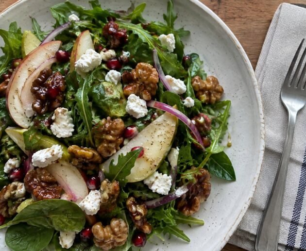 Ensalada de otoño con arúgula, pera en rebanadas, queso de cabra, nueces caramelizadas y granada en plato de cerámica sobre mesa de madera