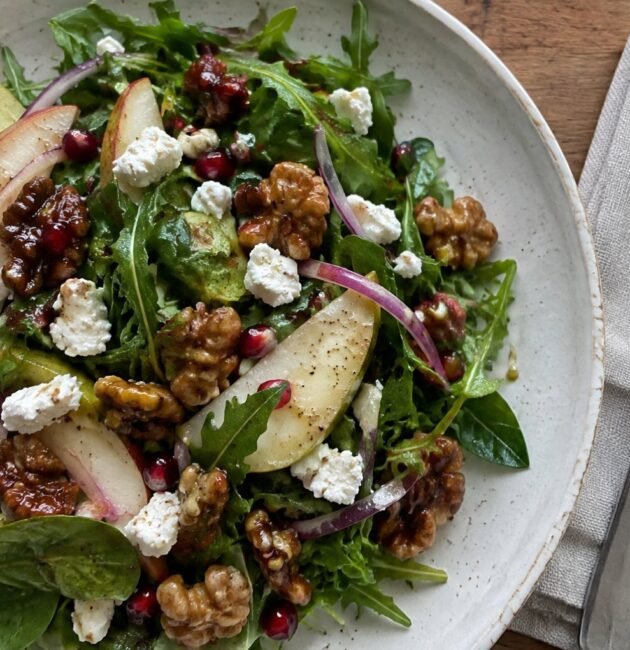 Ensalada de otoño con arúgula, pera en rebanadas, queso de cabra, nueces caramelizadas y granada en plato de cerámica sobre mesa de madera