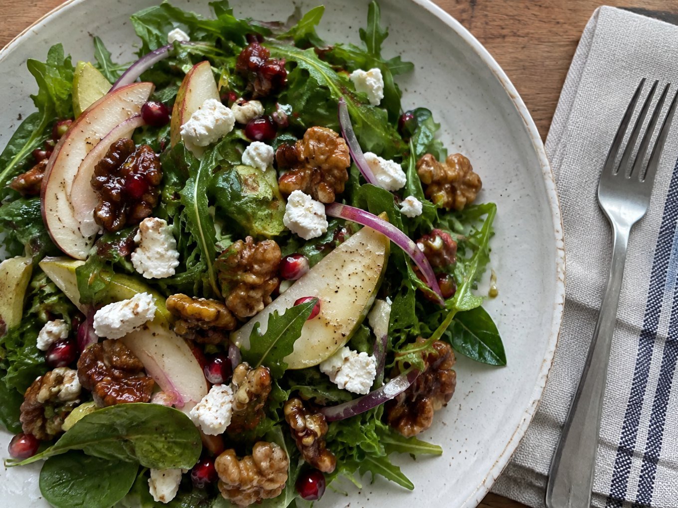 Ensalada de otoño con arúgula, pera en rebanadas, queso de cabra, nueces caramelizadas y granada en plato de cerámica sobre mesa de madera