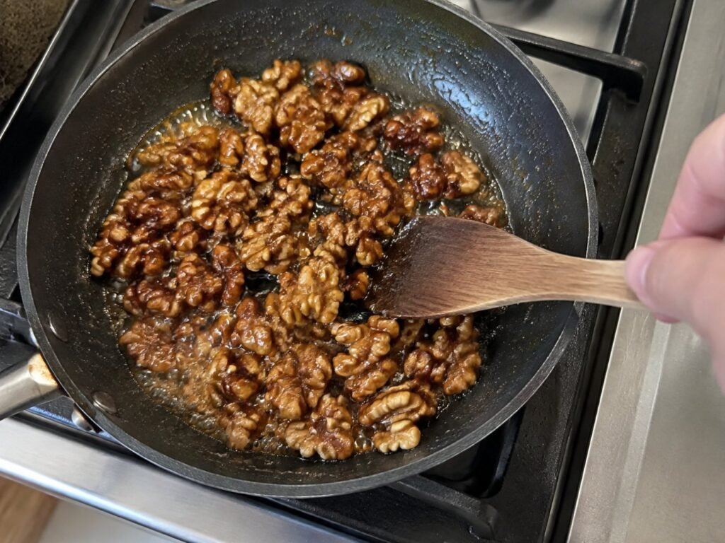 Mano moviendo nueces glaseadas con jarabe de maple en un sartén pequeño sobre la estufa, paso para hacer nueces caramelizadas caseras rápidas