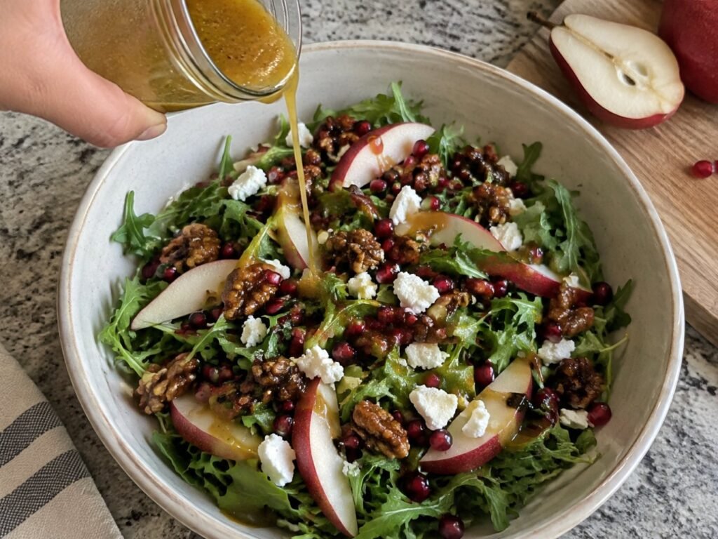 Mano vertiendo vinagreta dorada de mostaza Dijon y maple desde un frasco sobre ensalada de arúgula, pera y granada en un bowl grande de cocina