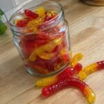 Frasco de vidrio con gomitas caseras de fresa y mango en forma de gusanitos hechas con agar-agar sobre una barra de madera
