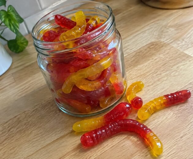 Frasco de vidrio con gomitas caseras de fresa y mango en forma de gusanitos hechas con agar-agar sobre una barra de madera