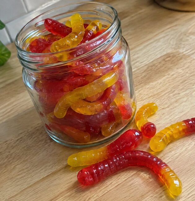 Frasco de vidrio con gomitas caseras de fresa y mango en forma de gusanitos hechas con agar-agar sobre una barra de madera