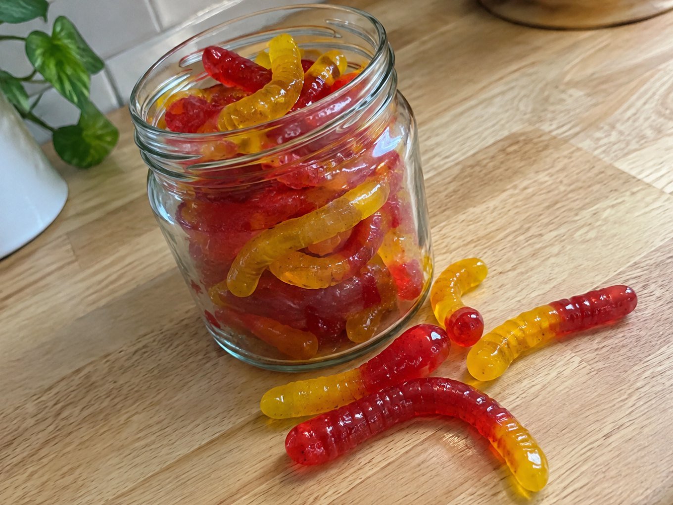 Frasco de vidrio con gomitas caseras de fresa y mango en forma de gusanitos hechas con agar-agar sobre una barra de madera
