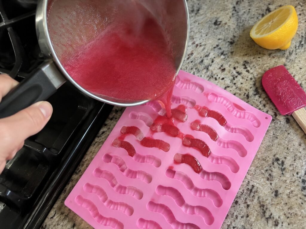 Mano vertiendo la mezcla caliente de fresa con agar-agar desde una olla pequeña a un molde de silicón de gusanitos en la cocina