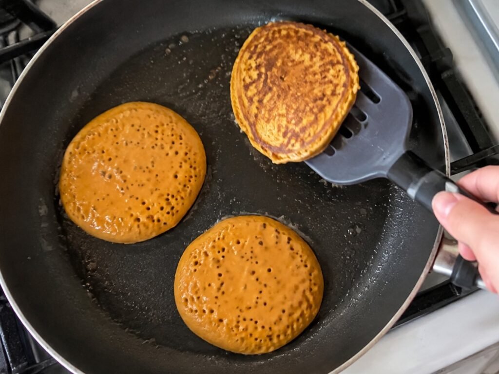 Mano volteando hot cakes de calabaza y avena en un sartén antiadherente, paso a paso de la receta de desayuno fácil de Halloween
