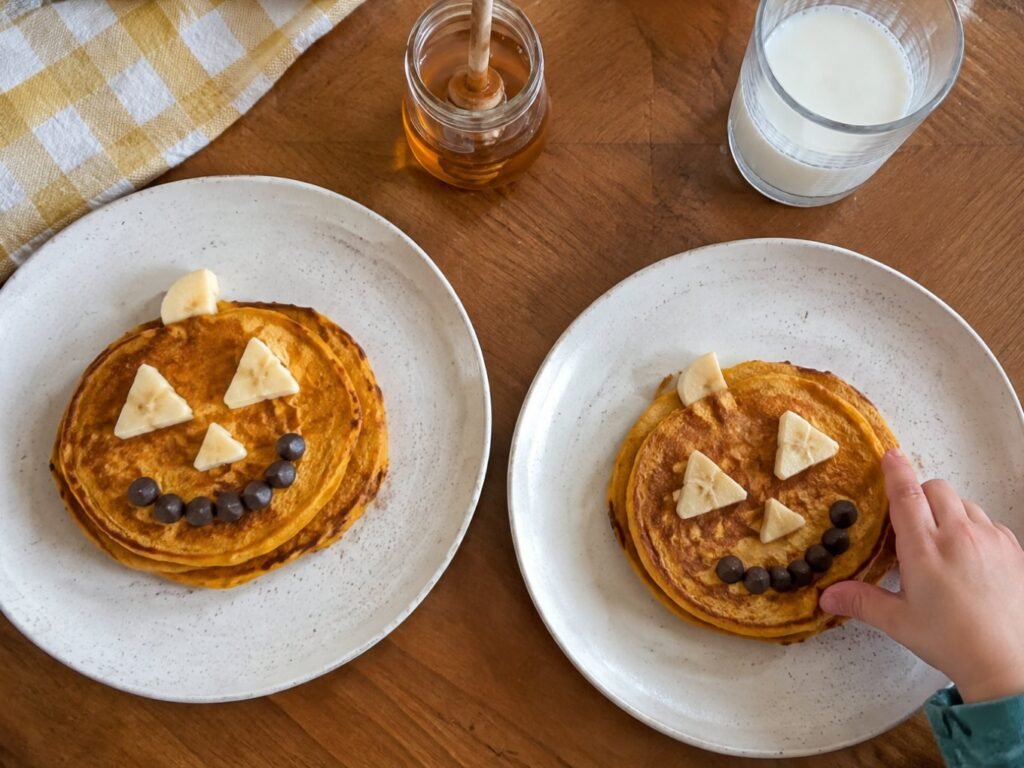Mesa de desayuno de domingo con platos de hot cakes de calabaza con caritas de Halloween, miel y leche, receta saludable para niños