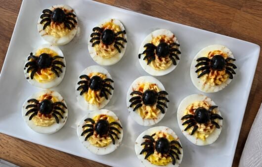 Charola de huevos rellenos araña con aceitunas negras y paprika, botana de Halloween fácil y alta en proteína sobre barra de cocina