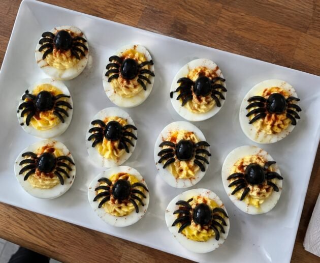 Charola de huevos rellenos araña con aceitunas negras y paprika, botana de Halloween fácil y alta en proteína sobre barra de cocina