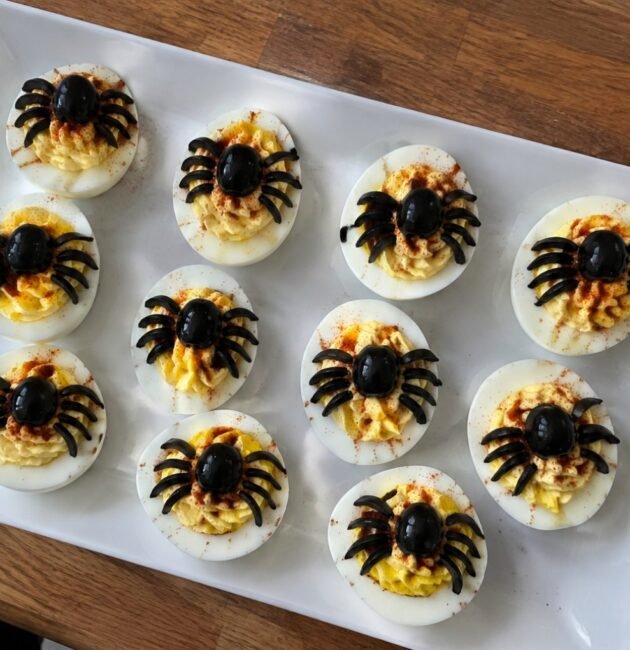 Charola de huevos rellenos araña con aceitunas negras y paprika, botana de Halloween fácil y alta en proteína sobre barra de cocina