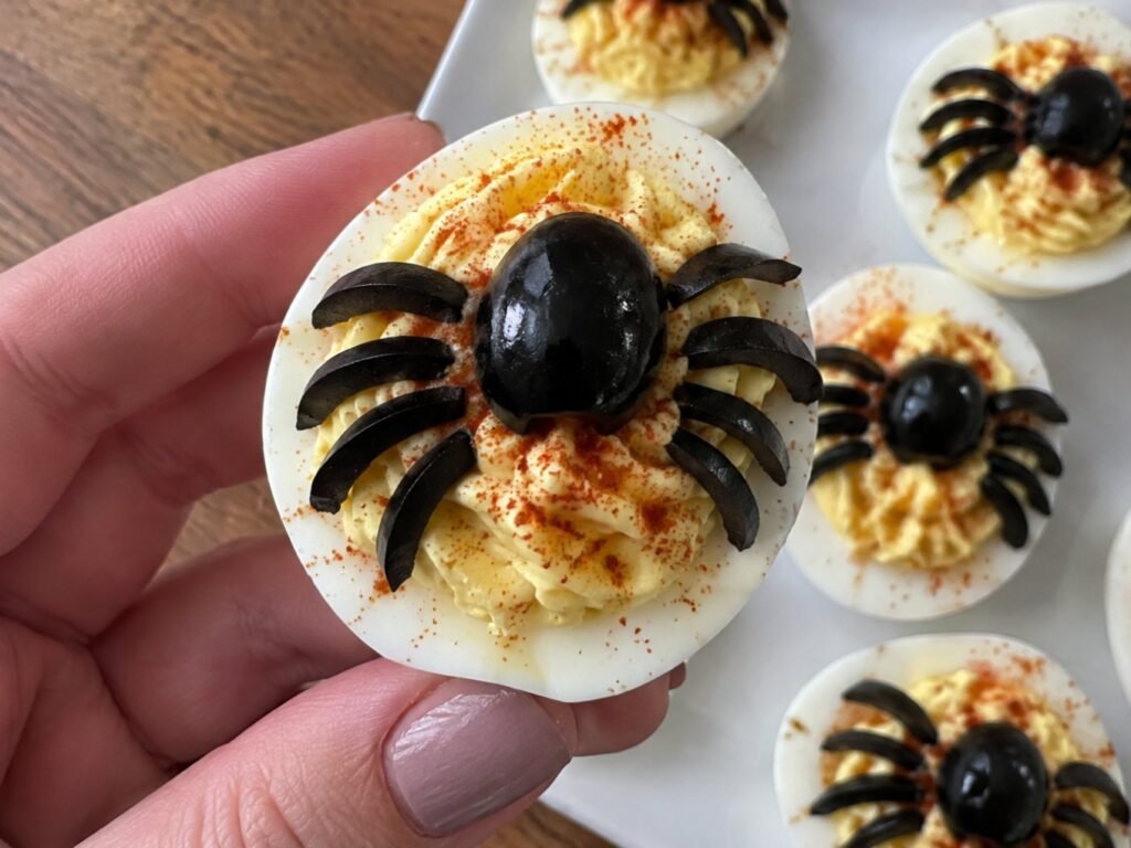 Mano sosteniendo un huevo relleno araña con relleno cremoso de yogur, paprika y patas de aceituna negra, botana saludable de Halloween