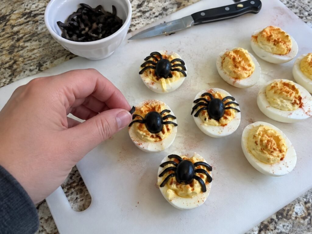 Mano colocando tiras de aceituna negra como patas de araña sobre huevos rellenos en una tabla de cocina, receta fácil de Halloween