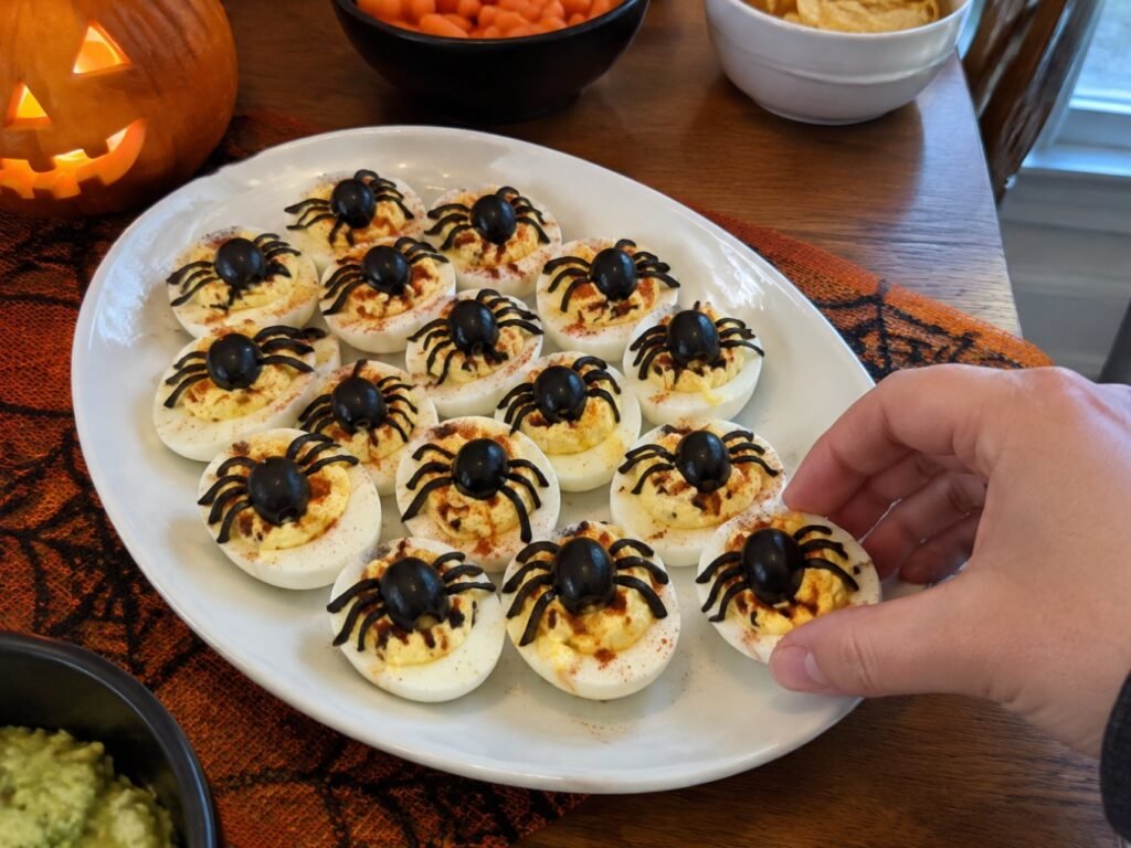 Charola de huevos rellenos araña en mesa de fiesta de Halloween con calabaza, mano tomando uno, botana salada para compartir