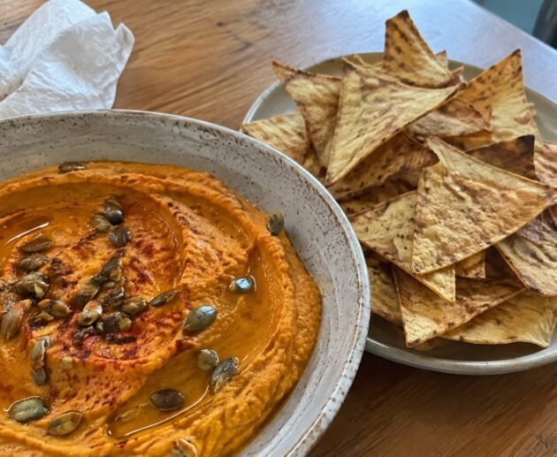 Hummus de calabaza asada naranja con aceite de oliva, paprika y pepitas, servido con chips de tortilla horneados en mesa de madera