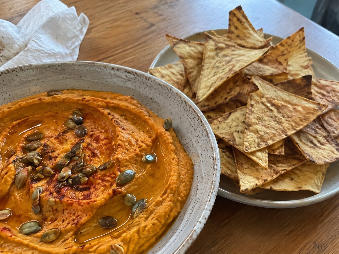 Hummus de calabaza asada naranja con aceite de oliva, paprika y pepitas, servido con chips de tortilla horneados en mesa de madera