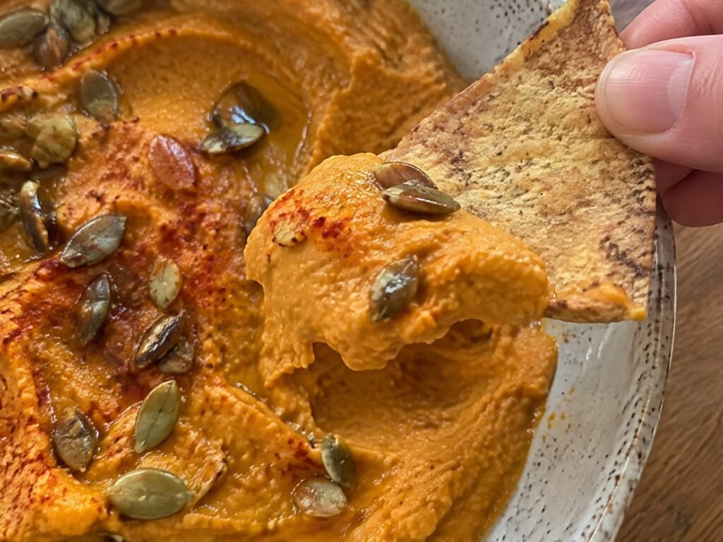 Mano sumergiendo un chip de tortilla horneado y crujiente en hummus de calabaza asada cremoso con semillas de calabaza tostadas