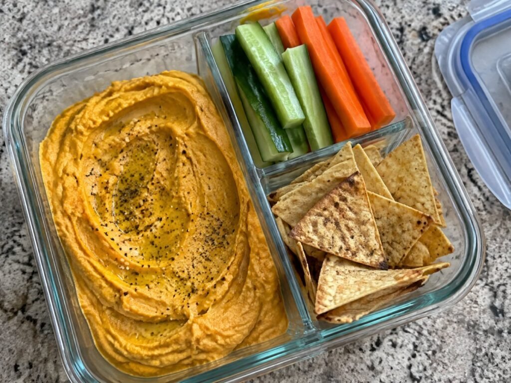 Recipiente de meal prep con hummus de calabaza asada, palitos de pepino y zanahoria y chips de tortilla horneados para llevar