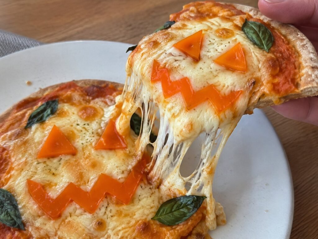 Mano levantando una rebanada de mini pizza de calabaza con mozzarella derretida que se estira y carita de pimiento naranja de Halloween