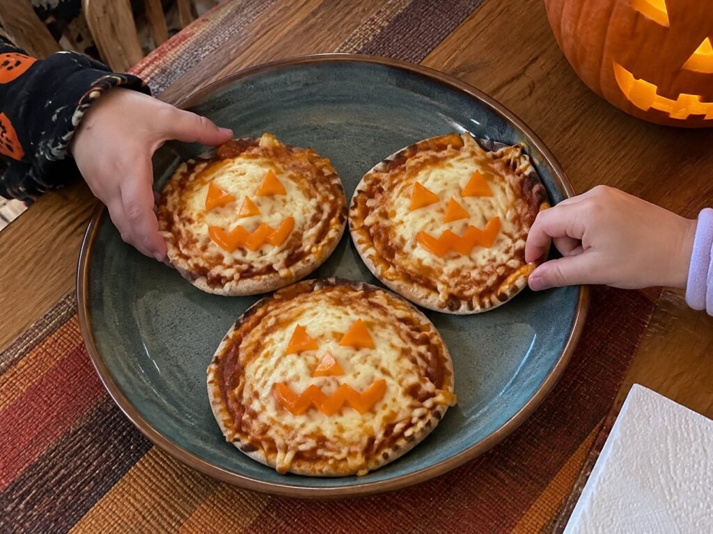 Manos de niños tomando mini pizzas de calabaza con caritas de pimiento naranja en una mesa casera de cena de Halloween con calabaza tallada