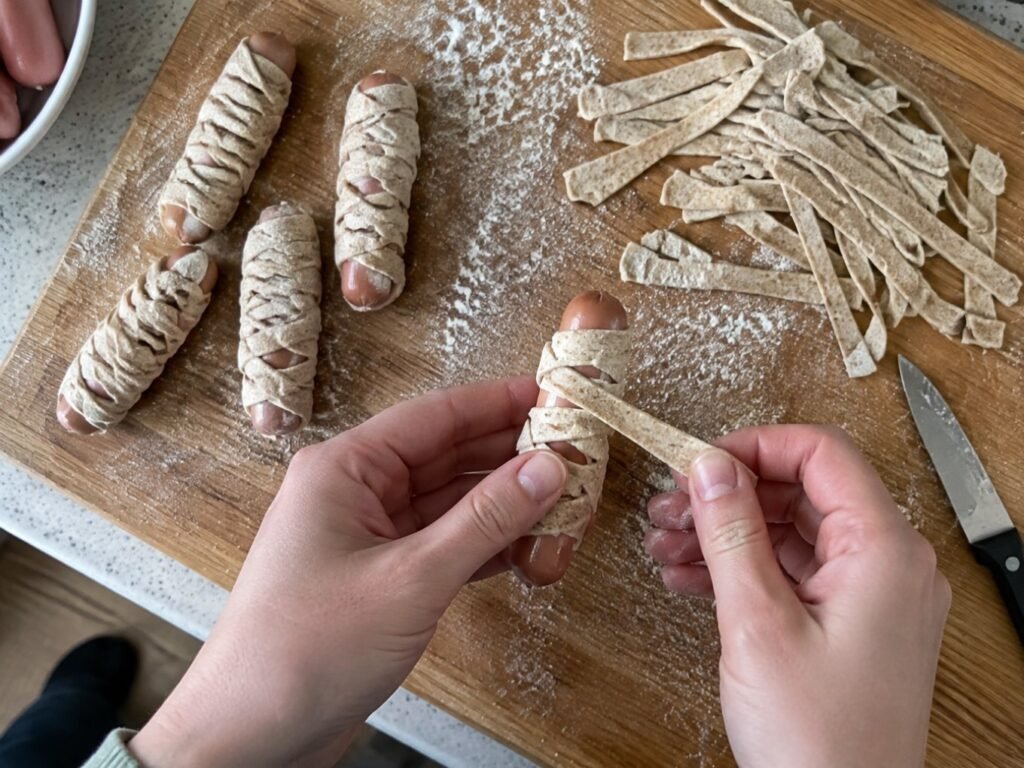 Manos enrollando tiras de masa integral casera alrededor de salchichas de pavo en tabla enharinada para hacer momias de Halloween
