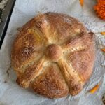 Pan de muerto integral casero con huesitos y azúcar sobre papel para hornear en la barra de la cocina, con pétalos de cempasúchil