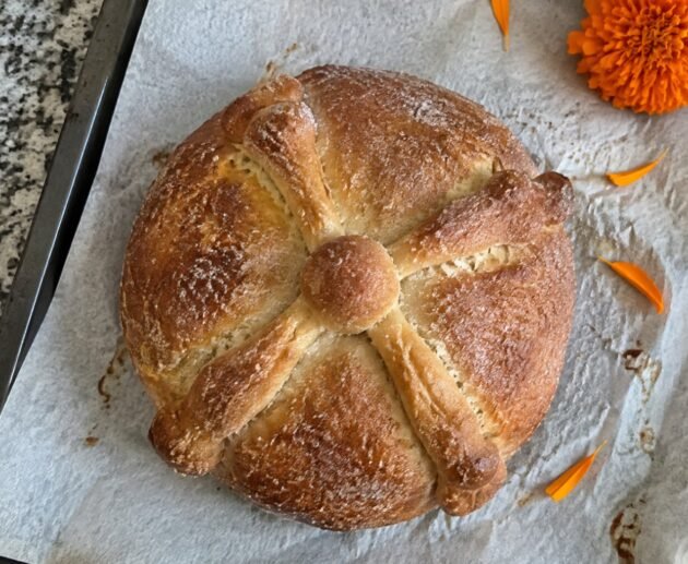 Pan de muerto integral casero con huesitos y azúcar sobre papel para hornear en la barra de la cocina, con pétalos de cempasúchil