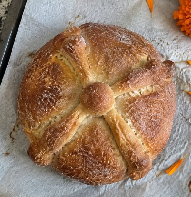 Pan de muerto integral casero con huesitos y azúcar sobre papel para hornear en la barra de la cocina, con pétalos de cempasúchil