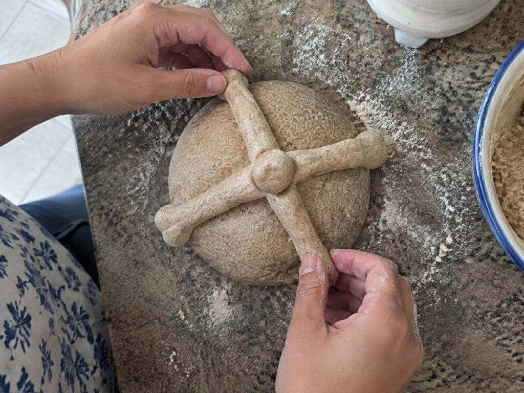 Manos formando y colocando los huesitos de masa sobre una bola de masa integral para pan de muerto en la barra enharinada