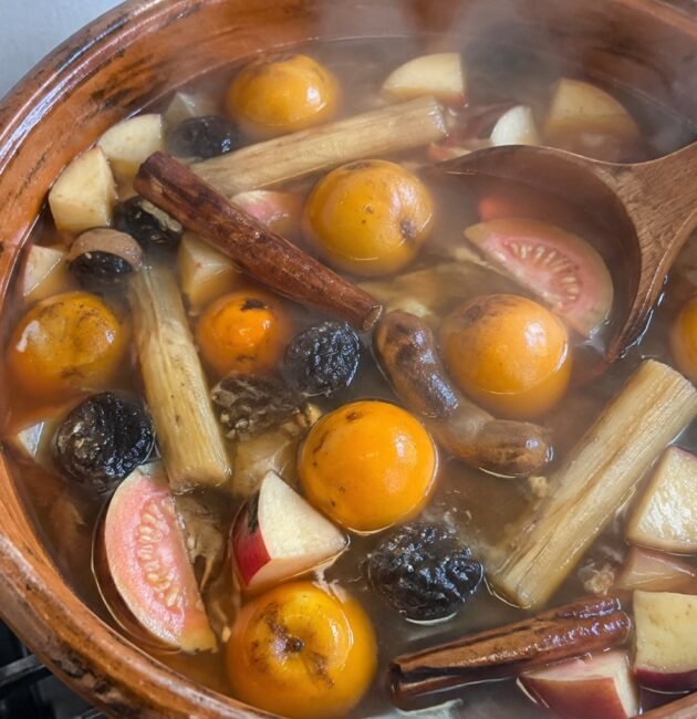 Olla de barro con ponche navideño de frutas sin azúcar humeante, con tejocotes, guayaba, caña y canela sobre la estufa de casa