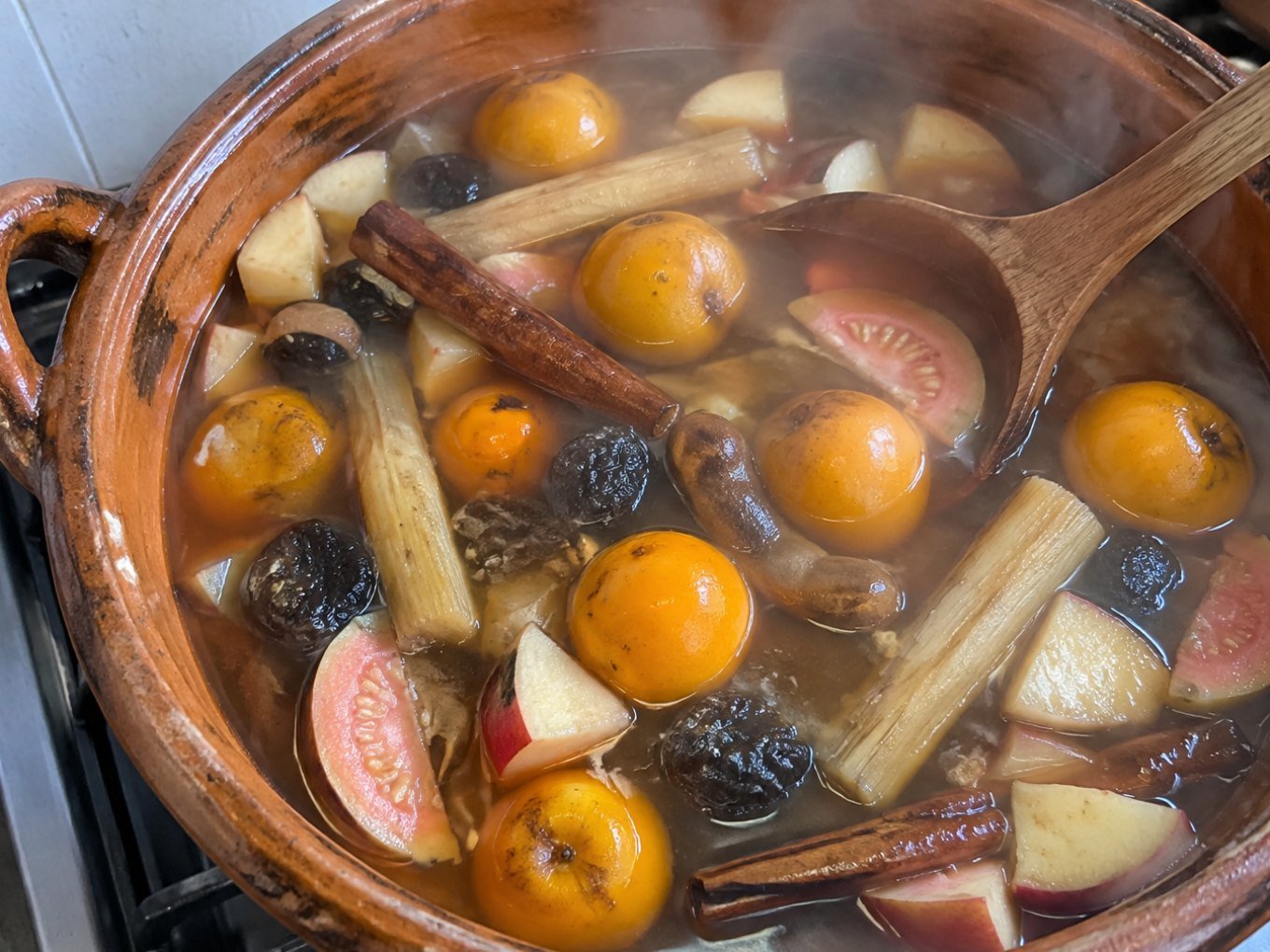 Olla de barro con ponche navideño de frutas sin azúcar humeante, con tejocotes, guayaba, caña y canela sobre la estufa de casa