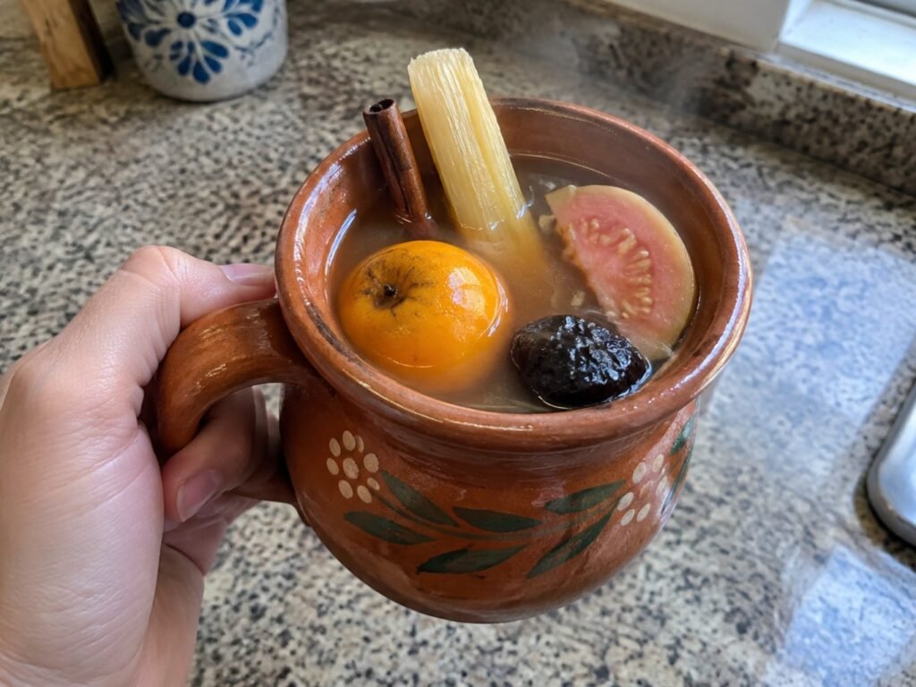 Mano sosteniendo un jarrito de ponche navideño caliente con bastón de caña y tejocote, ponche de frutas sin azúcar añadida en casa